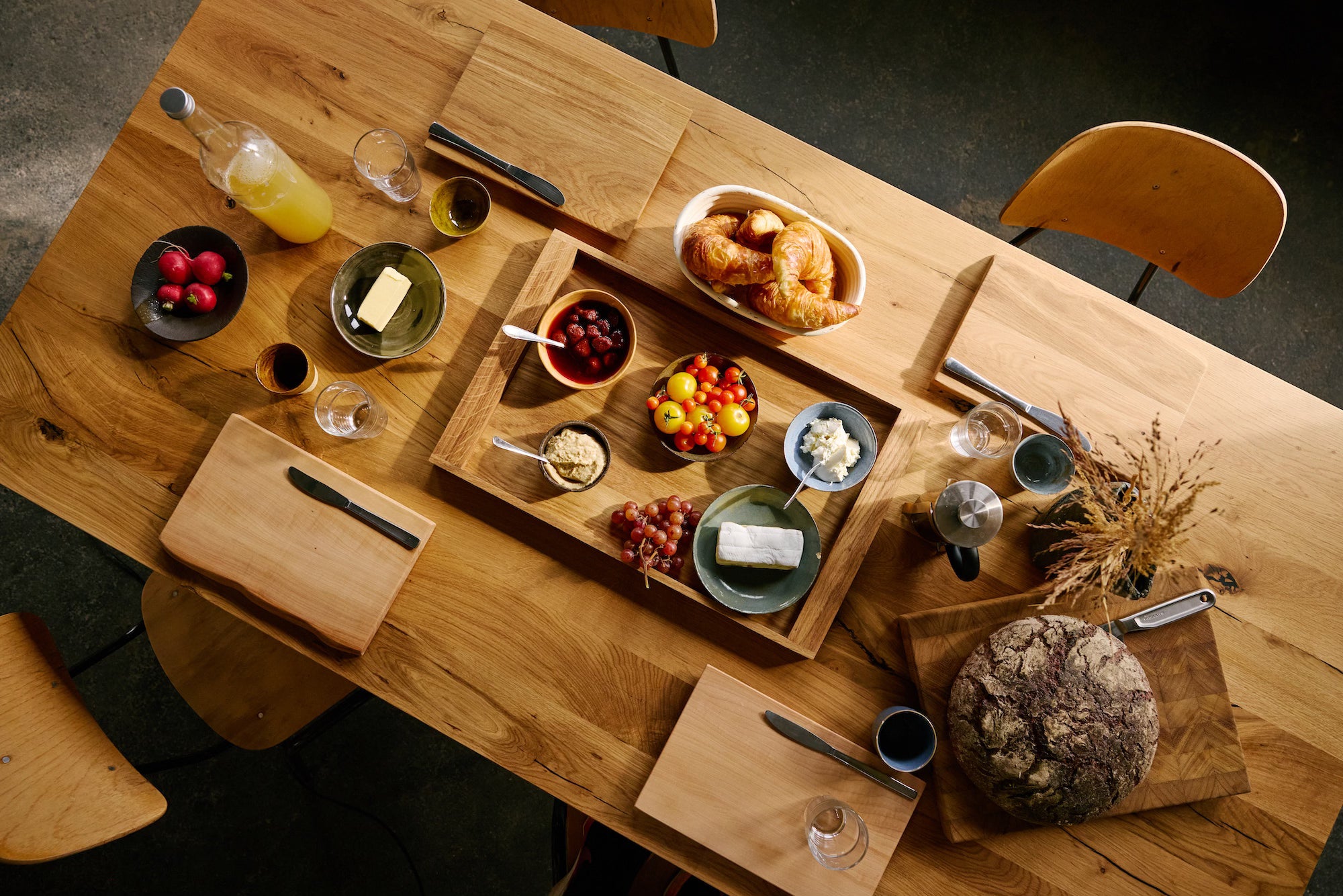 Massivholztisch mit Schneidebrettern, Croissants, Brot, Frühstücksschalen und Holzstühlen.
