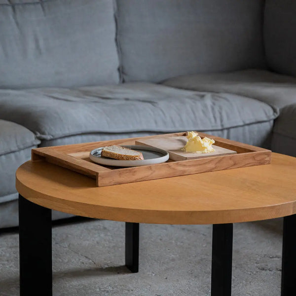 Minimalistisches Serviertablett aus Eiche auf Holztisch. Darauf ein Teller mit Brot und ein Holzbrett mit Butter. Graues Sofa im Hintergrund.