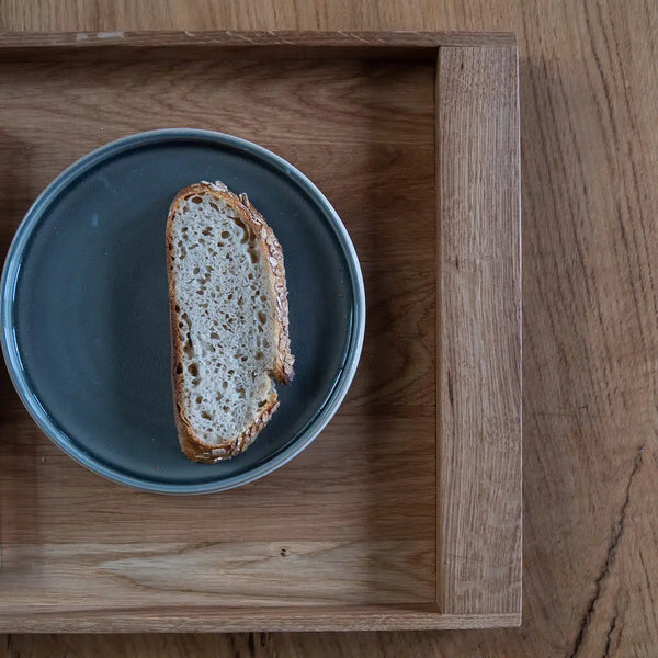 Serviertablett aus Eiche mit blauem Teller und Brot. Holzstruktur sichtbar. Nachhaltiges, minimalistisches Design.